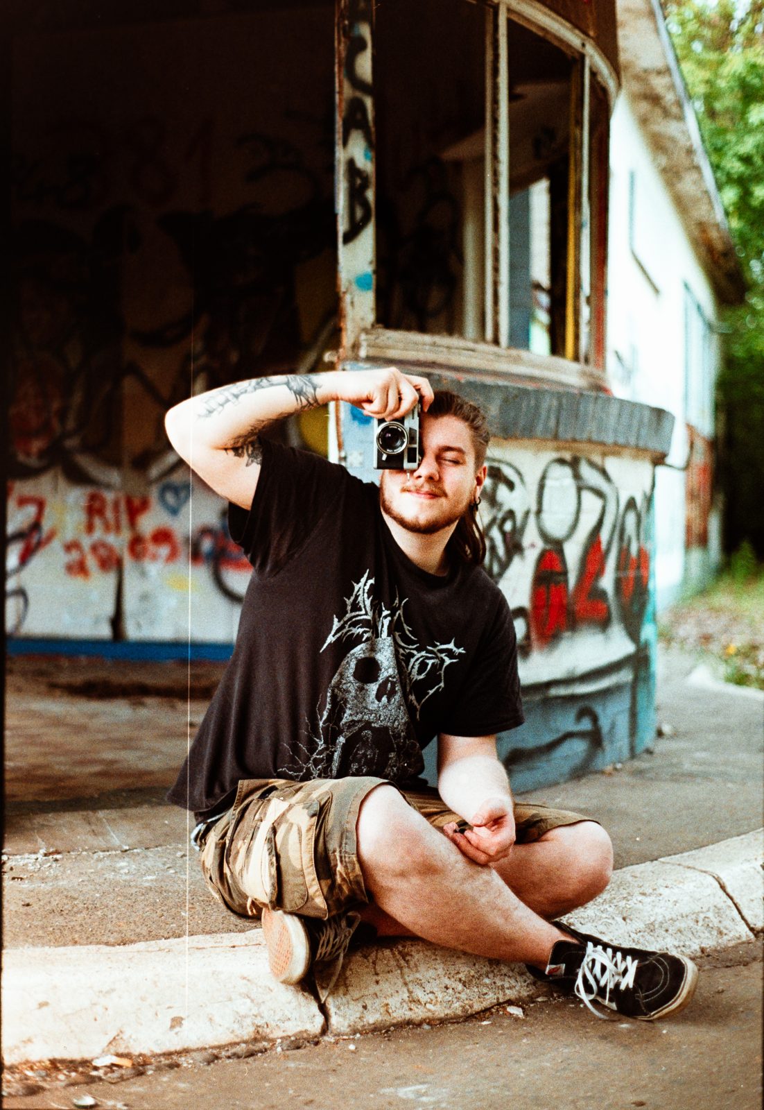Jan sitzt vor einer Graffiti-Wand und fotografiert mit einer Analogkamera